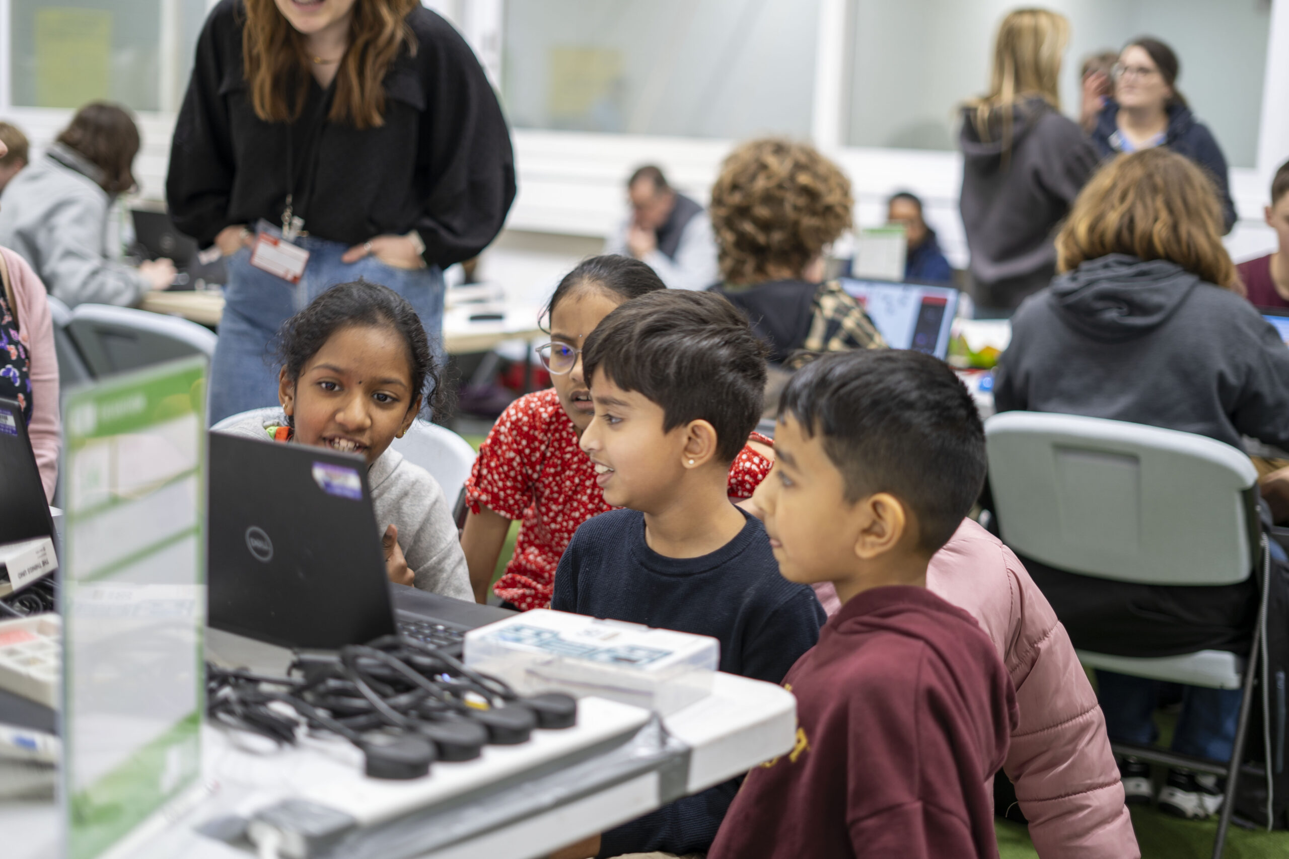 Four children with happy faces huddled behind a laptop in the busy Techathon competition room. Beside their laptop is a electronics maker kit and many wires. Behind the children, other competitors are seen working hard on their projects.
